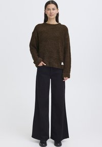 Maglione marrone e peloso con scollo rotondo e maniche lunghe, abbinato a pantaloni neri a vita alta e a gamba larga. Etichetta del marchio visibile e discreta.