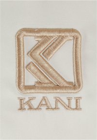 Brodert logo med et geometrisk design i gulltråd på en hvit stoffbakgrunn, med bokstavene "KANI" under designet.