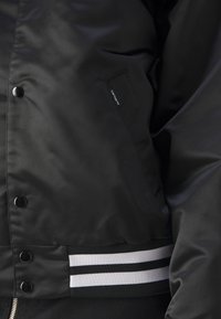 Schwarze Bomberjacke mit glatter Außenseite, Druckknöpfen, seitlichen Taschen und gestreiften Rippbündchen in Schwarz und Weiß.