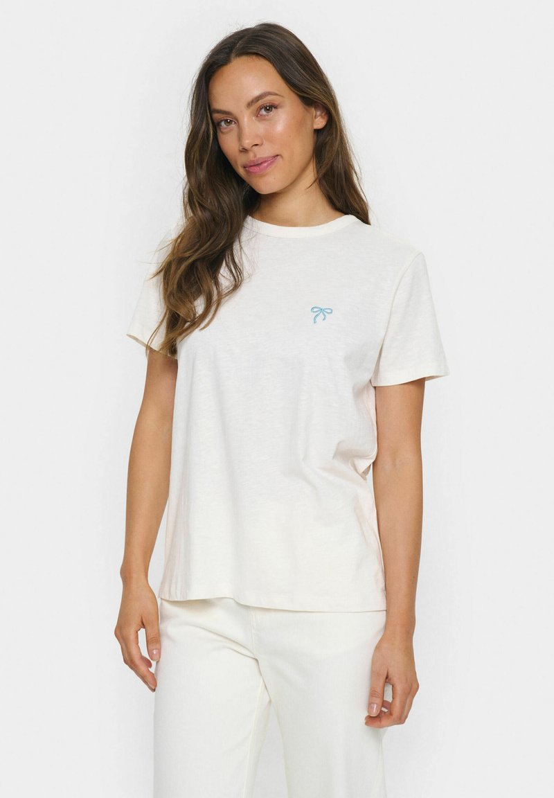 Camiseta de manga corta color crema con cuello redondo y un lazo azul claro bordado en el lado izquierdo. Fabricada con un tejido suave.