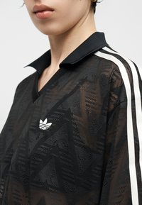 Άτομο που φοράει μαύρο διαπνέον μπλουζάκι Adidas με λευκές ρίγες στον ώμο και λευκό λογότυπο Adidas στο στήθος, κοντινό πλάνο στο άνω μέρος του κορμού.