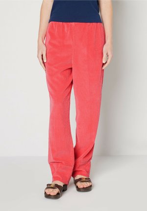 Persoon draagt loszittende koraalkleurige corduroy broek met elastische tailleband en donkerbruine open sandalen, staand tegen een effen achtergrond.