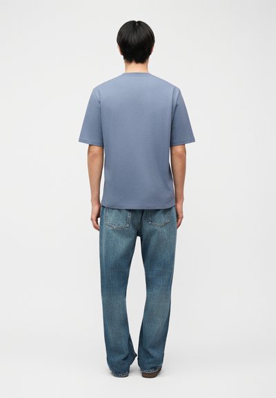 Άνδρας που στέκεται με την πλάτη προς τα έξω φορώντας απλό μπλε μπλουζάκι με κοντά μανίκια και χαλαρά μπλε τζιν παντελόνια μπροστά σε απλό φόντο.