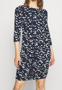 Mörkblå klänning med trekvartslånga ärmar, prydd med ett blommönster i vitt och ljusrosa. Rynkad textur förbättrar formen.