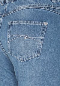 Primer plano de un bolsillo trasero de unos jeans de mezclilla azul con detalle de costura en zigzag blanca y remache de metal en la esquina superior derecha.