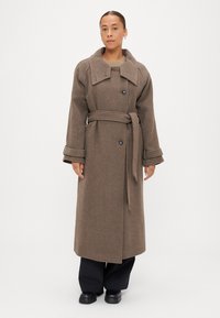 Brūns vilnas mētelis ar lielu apkakli, dubultām pogām, jostu viduks un teksturēta auduma, kombinēts ar melnām biksēm un apaviem.