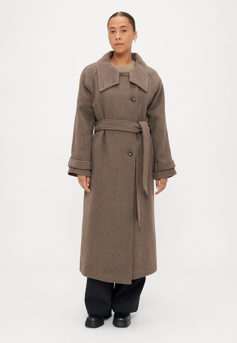 Brūns vilnas mētelis ar lielu apkakli, dubultām pogām, jostu viduks un teksturēta auduma, kombinēts ar melnām biksēm un apaviem.