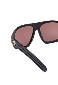 Occhiali da sole con montatura nera, dotati di grandi lenti marroni, un design elegante e un'accortezza dorata sulla tempia con un dettaglio del logo.