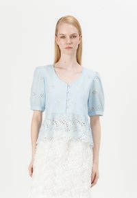 Blusa azzurro chiaro con maniche a palloncino, chiusura con bottoni e ricami floreali. L'orlo inferiore presenta un sovrapposto in pizzo bianco con motivi intricati.