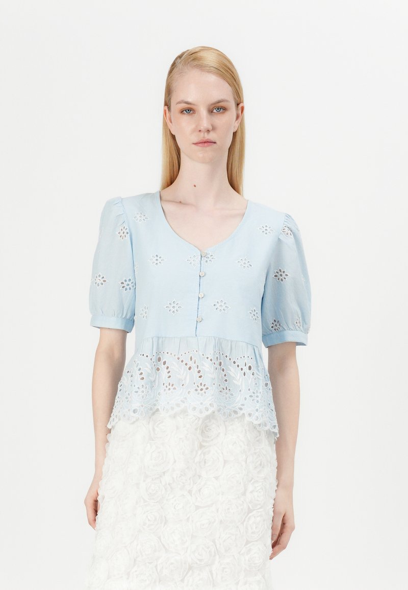 Blusa azzurro chiaro con maniche a palloncino, chiusura con bottoni e ricami floreali. L'orlo inferiore presenta un sovrapposto in pizzo bianco con motivi intricati.