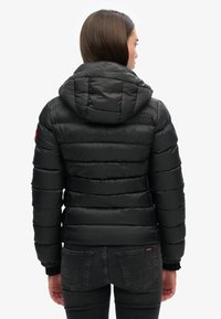 Zwarte pufferjack met een capuchon, voorzien van een gevoerde textuur en elastische manchetten. Zichtbaar logo op de schouder en een stijlvolle pasvorm.