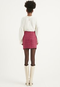 Top lungo bianco texturizzato con scollatura a volant, abbinato a una gonna a scacchi rossa aderente e stivali beige fino al ginocchio.
