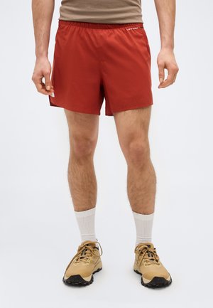 Man draagt roestkleurige sportshorts, beige sneakers en witte crew-sokken tegen een effen lichte achtergrond.