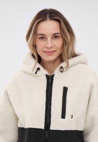 Fleece hooded jacket with cream top half and black bottom. Features a front zipper and a small pocket with contrasting black detail.