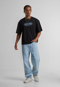 Tricou oversized din bumbac negru cu logo-ul albastru "KANI", asortat cu blugi lejeri de un albastru deschis și adidași albi. Accesoriu simplu dintr-un lanț de argint.