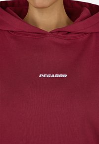 Burgundská mikina s kapucňou obsahujúca predné klokanie vrecko, šnúrku na kapucni a biele vyšívané logo "PEGADOR" na hrudi. Mäkká, hladká textúra.