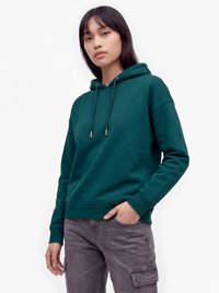 Junge Frau, die einen dunkelgrünen Hoodie und graue Cargohosen trägt, steht mit einer Hand in der Tasche vor einem schlichten weißen Hintergrund.