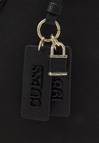 Sac à main texturé noir avec deux étiquettes : l'une avec "GUESS" gaufré et l'autre avec "1981", agrémenté de ferrures et d'un porte-clés dorés.