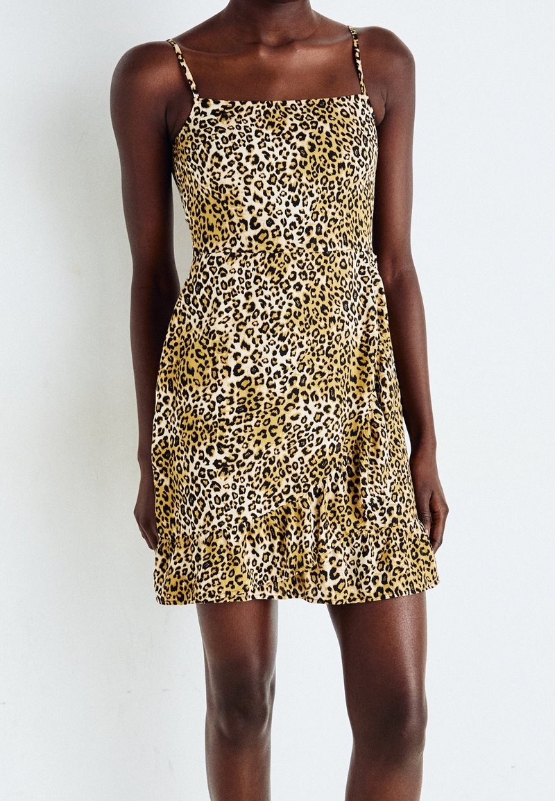 Femme noire portant une mini-robe sans manches à imprimé léopard avec de fines bretelles, debout devant un fond blanc uni.