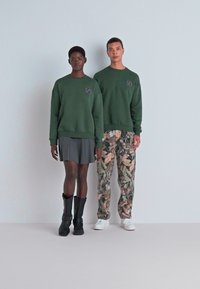 Felpe verdi con logo, gonna grigia e stivali neri su un modello; pantaloni camouflage e scarpe bianche su un altro modello. Sfondo semplice.