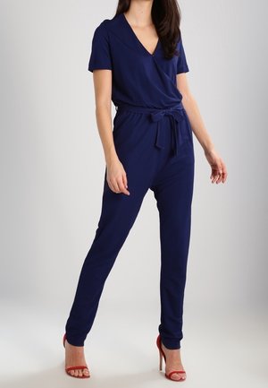 Granatowy kombinezon z dekoltem w serek i krótkimi rękawami, z paskiem w talii i zwężanymi nogawkami; wykonany z gładkiej, lekkiej tkaniny.