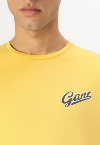 Gelbes Baumwoll-T-Shirt mit Rundhalsausschnitt und einem blauen, geschwungenen "Gant"-Logo auf der linken Brustseite. Glatte Textur und klassischer Schnitt.
