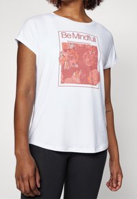 Biała koszulka z krótkim rękawem z różowym kwiatowym wzorem z tekstem w prostokątnej ramce. Materiał wydaje się miękki i lekki.