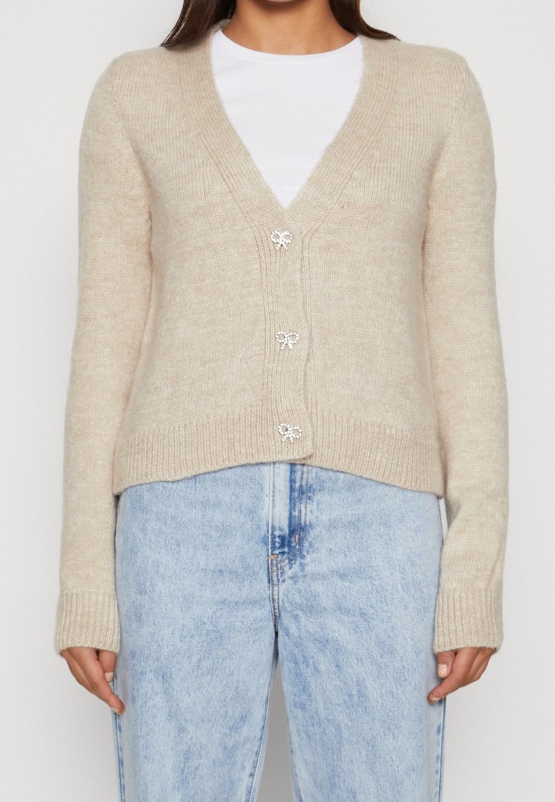 Cardigan beige en tricot avec col en V, orné de trois boutons décoratifs avec nœuds, et poignets côtelés, associé à un jean bleu clair.
