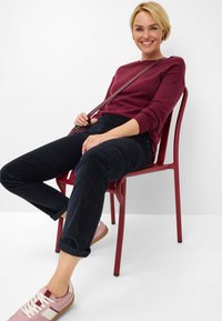 Bordeaux trui, donkerblauwe broek met geplooide details en roze sneakers met witte accenten. Zittend op een rode stoel tegen een witte achtergrond.