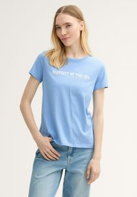 Camiseta de algodón azul claro con la inscripción "HAPPIEST BY THE SEA" en blanco, de manga corta, cuello redondo y un corte relajado.