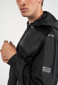 Giacca nera con un pannello frontale lucido, polsini elasticizzati e cappuccio. Include una patch del logo sulla manica sinistra.