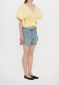 Gelbe, kurzärmelige Bluse mit tiefem V-Ausschnitt und gerafftem Design, kombiniert mit abgetragenen Jeans-Shorts und schwarzen Ballerinas.