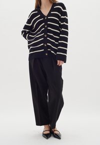 Žena v černobíle pruhovaném cardiganu s knoflíky, volných černých kalhotách a černých špičatých balerínách stojící proti jednobarevnému pozadí.