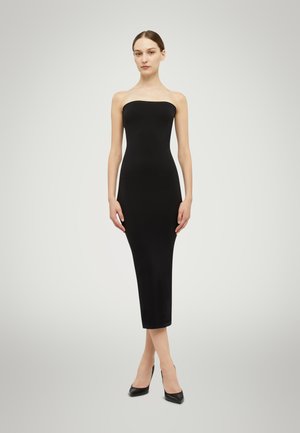 Zwarte, strapless, nauwsluitende jurk van gladde stof, tot halverwege de kuit reikt, in combinatie met puntige zwarte hakken, op een licht vervaagd achtergrond.