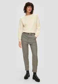 Vrouw met krullend haar, gekleed in een off-white langarmtop, zwart-wit houndstooth broek en zwarte platform enkellaarzen, staande met één hand in haar zak.