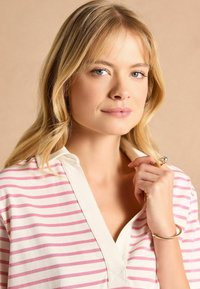 Balta un rozā svītrainā blūze ar apkaklītes izgriezumu, izgatavota no mīksta auduma, ar vieglu V formas izgriezumu un dekoratīvu zelta aproci uz modeļa rokas.