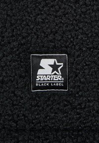 Stof de fleece negru cu o textură stratificată, având un etichetă albă rectangulară pe care este inscris logo-ul "STARTER" și textul "BLACK LABEL".