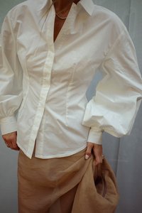 Witte blouse met knopen, pofmouwen, kraag en subtiele textuur, gecombineerd met een beige rok en met een bruine suède tas in de hand.