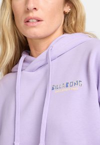 Sudadera morada hecha de tela suave, con capucha ajustable y un logo gráfico en azul y amarillo que dice "BILLA BONG SINCE SEVENTY THREE."