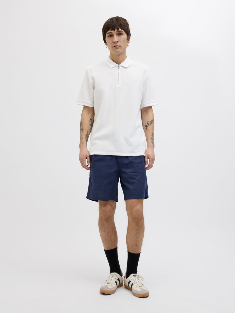 Witte polo met korte mouwen en een ritssluiting, gecombineerd met marineblauwe shorts. Sneakers hebben beige en groene accenten.