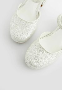 Pantofi pentru copii strălucitori din argint cu un design cu vârful rotund, având o curea albă și detalii perforate delicate pe margine.