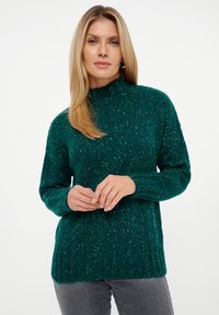 Zeleni pleteni pulover s visokim ovratnikom, rebrastim rubom i dugim rukavima. Tekstura sadrži točkice svjetlijih boja kroz cijeli materijal.