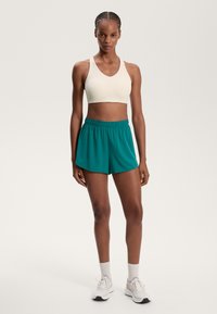 Helle beige sports-bh, kombinert med teal shorts med elastisk midje. Modellen har på seg hvite joggesko og rillete sokker.