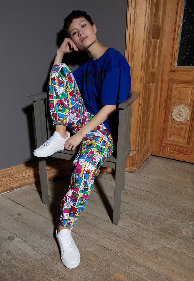 Camiseta azul oversized, leggings coloridos com padrão de quadrinhos e ténis brancos. Sentada numa cadeira de madeira contra uma parede cinza.