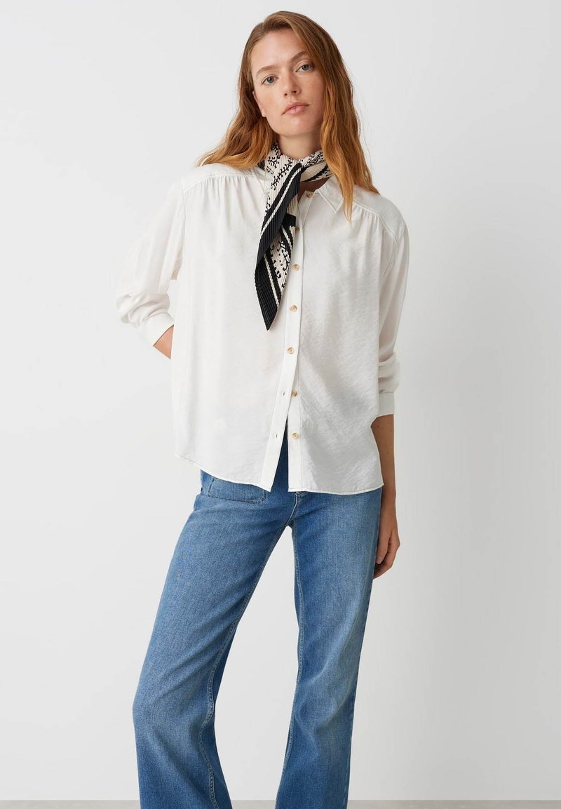 Camicia bianca con bottone, dal taglio rilassato, maniche lunghe e una sciarpa a motivi bianco e nero annodata intorno al collo, abbinata a jeans blu.