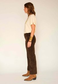 Chemise beige à manches courtes, pantalons en velours côtelé marron, bracelet de poignet en or, bottes brunes à bout pointu. Profil latéral sur un fond neutre.