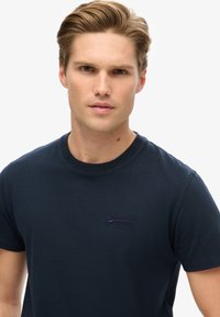 Ung mand med lysebrunt hår iført en marineblå Superdry t-shirt, vender fremad mod en ensfarvet hvid baggrund.