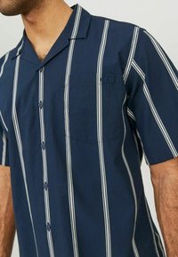 Tamsiai mėlyna trumparankovė marškiniai su baltais vertikaliais dryžiais, matiniai mygtukai, priekinė kišenė ir subtilus logotipo siuvinėjimas. Lengvos medžiagos tekstūra.