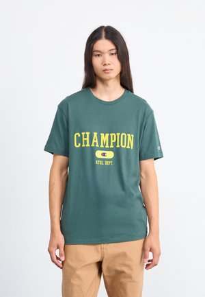 Ung person med langt hår bærer en grøn Champion T-shirt med gul skrift og beige bukser, stående foran en ensfarvet hvid baggrund.