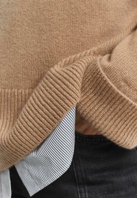 Un pull en tricot beige superposé sur une chemise blanche à fines rayures noires, associé à un jeans en denim noir.